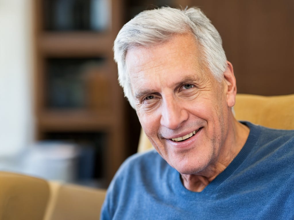White man with white hair and no glasses smiles, wearing a blue t-shirt, smiles into camera in this portrait photo.