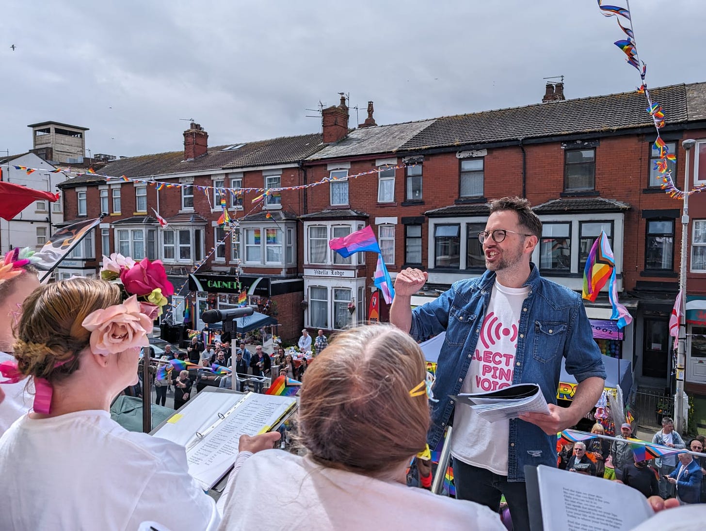 Phill Fairhurst conducting a choir on top of a bus in a crowded street at Blackpool Pride