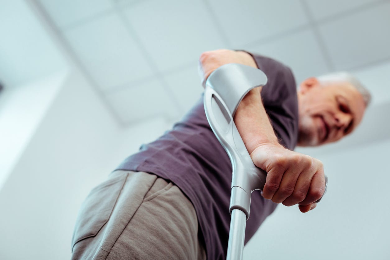 Camera looks up from the floor along right side of white man with gray hair. In foreground, the man’s fist grabs handle of Canadian crutch.  