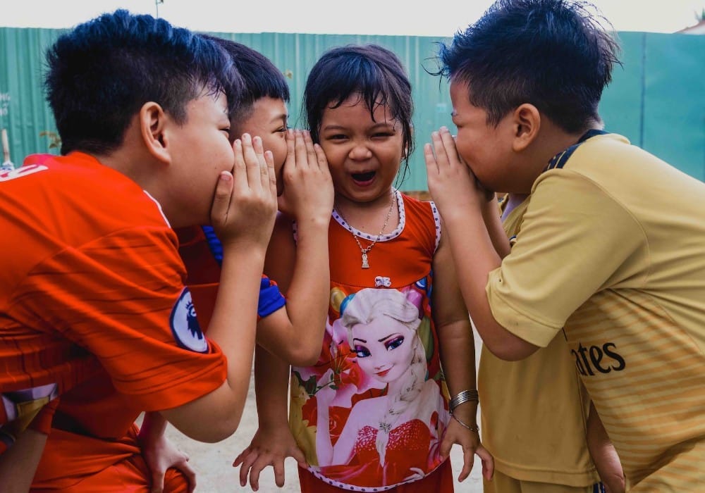 Group of Asian kids whispering to a girl who is laughing and making a funny face