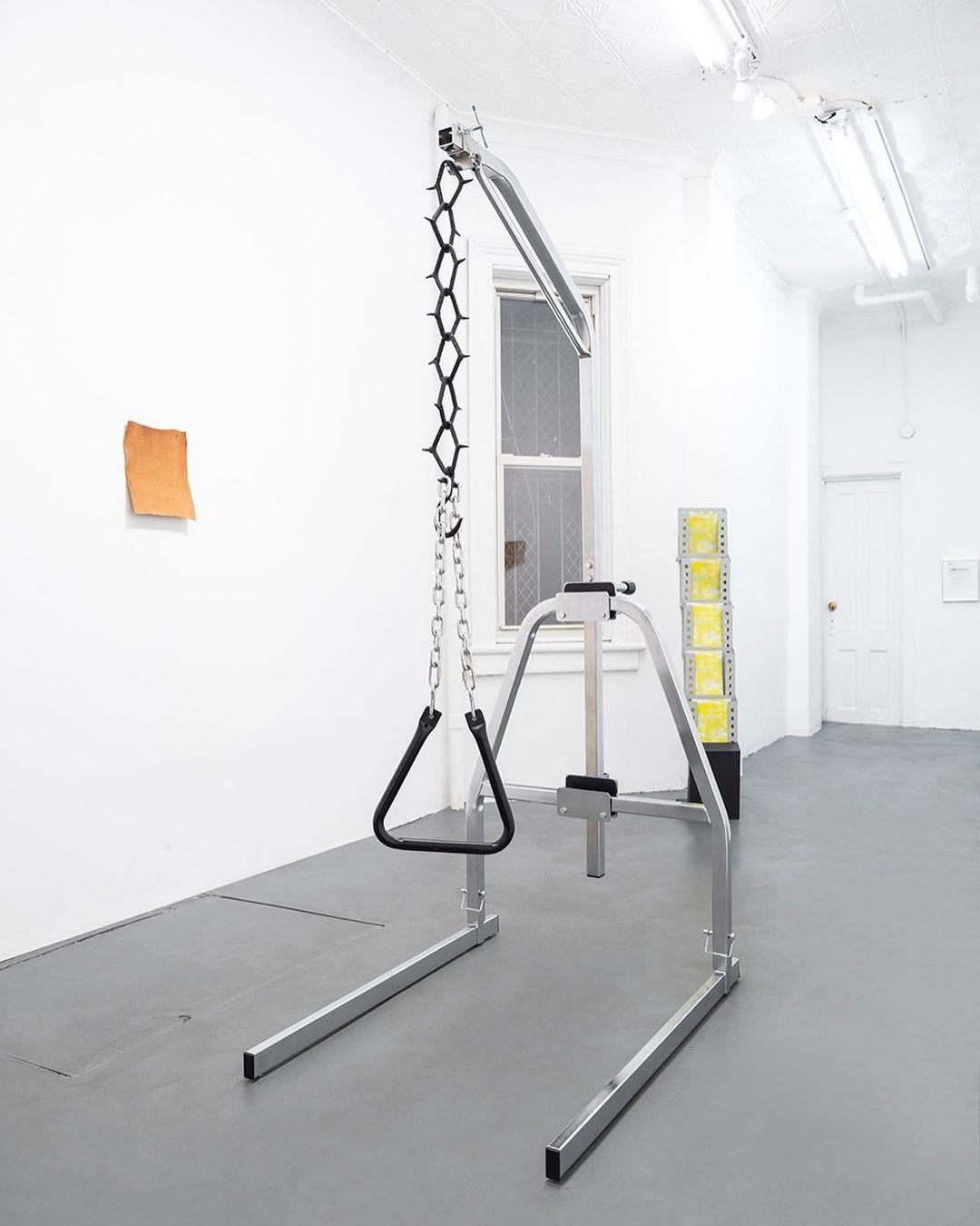 A metal trapeze handle is suspended from chains and nipple clamps within a bariatric trapeze frame that seems sturdy on the ground of a white-walled gallery.