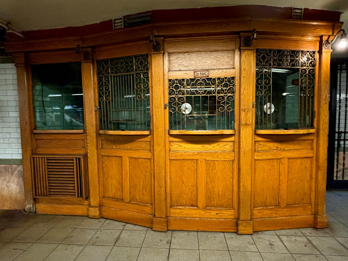 A wooden subway ticket booth. The three windows on the right push forward like half a hexagon. The metal grating protecting the person inside is ornate, bronze-tobed and curled. The left-most window is unadorned. The wood is warm and medium in shade. The inside of the booth is dark as no one is working at the moment. The center window has a slight reflection of the author's hands holding up her phone to take the picture. Over the window is small wood box inscribed R "204."