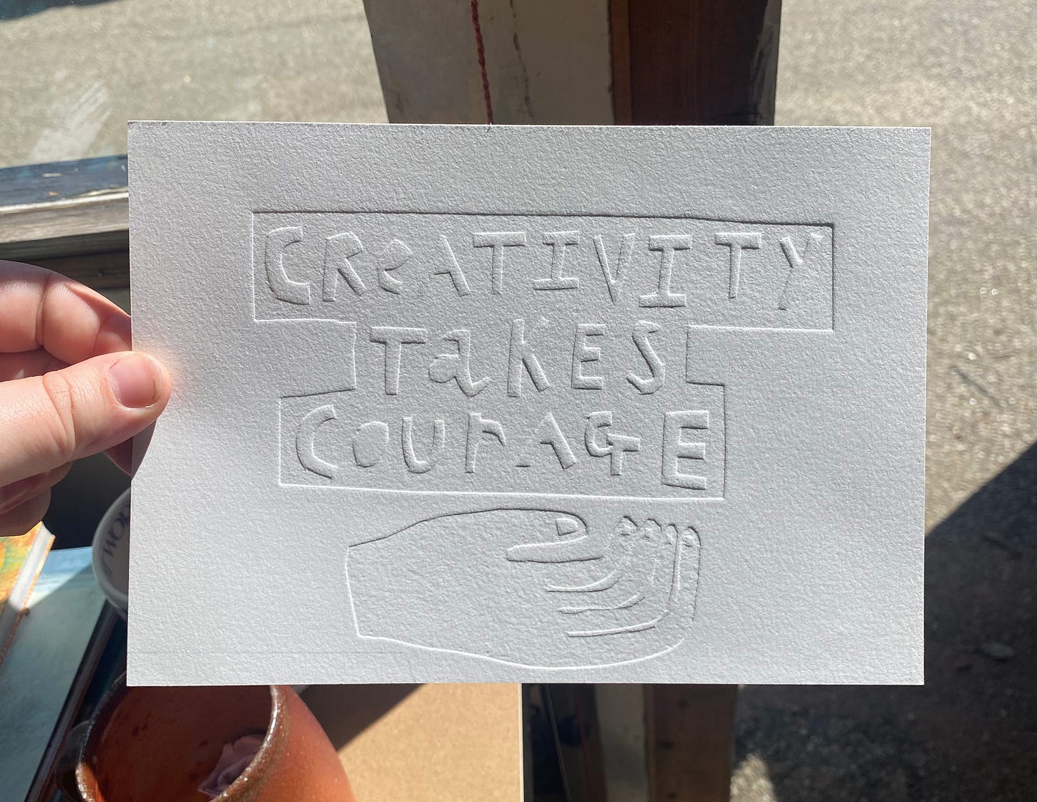 An embossed piece on white art paper which has the text creativity takes courage on and a styalised curled hand underneath.