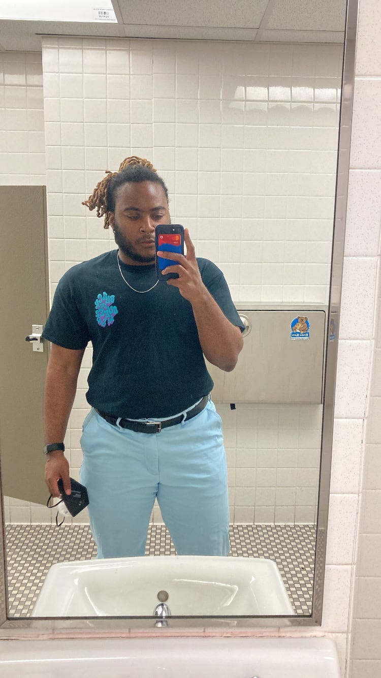 KB, a black person with black/blond locs, poses in front of a mirror in a bathroom. they are wearing blue pants and a black and blue shirt.
