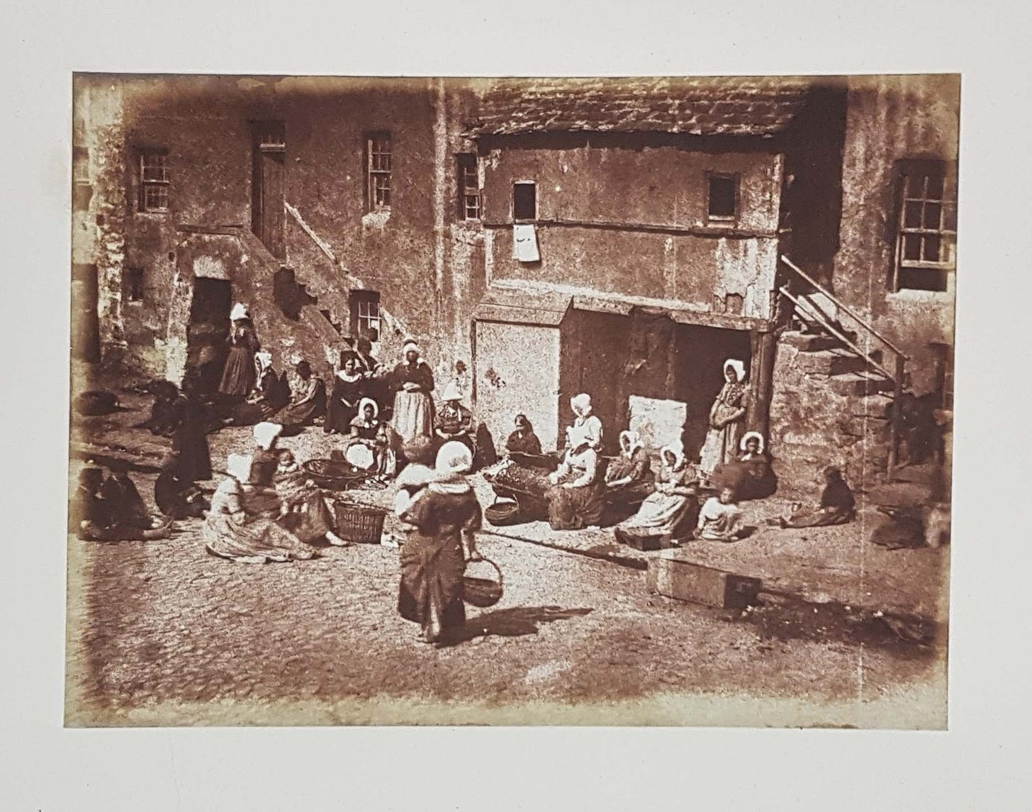 Photographic print showing women seated in front of houses preparing fishing lines. A woman holding a baby approaches them.