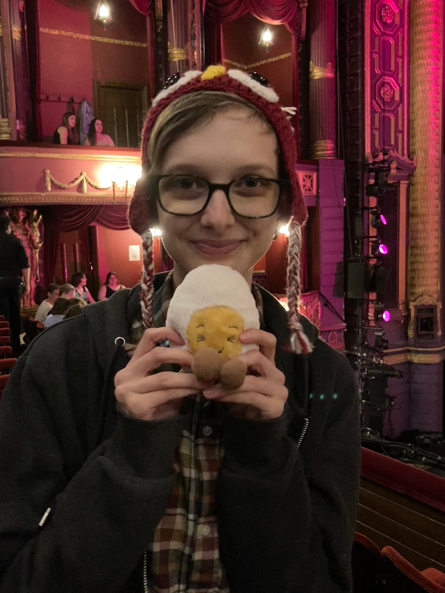 Photograph of Beck holding a soft toy boiled egg and smiling inside an ornate theatre. Beck is wearing an owl hat and glasses.