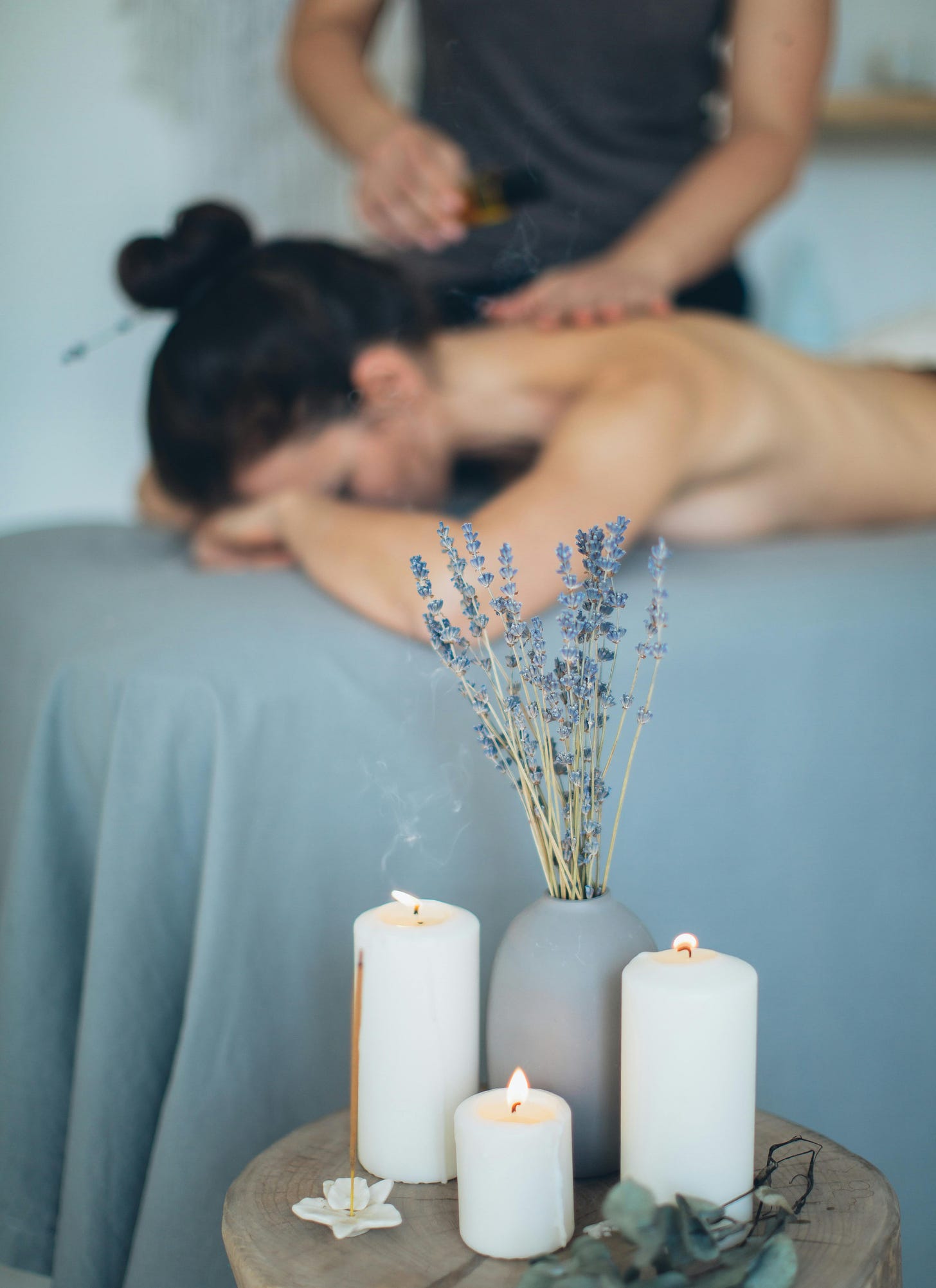 Woman lying face down on massage table with vase of lavendar and candles in foreground and masseuse in background