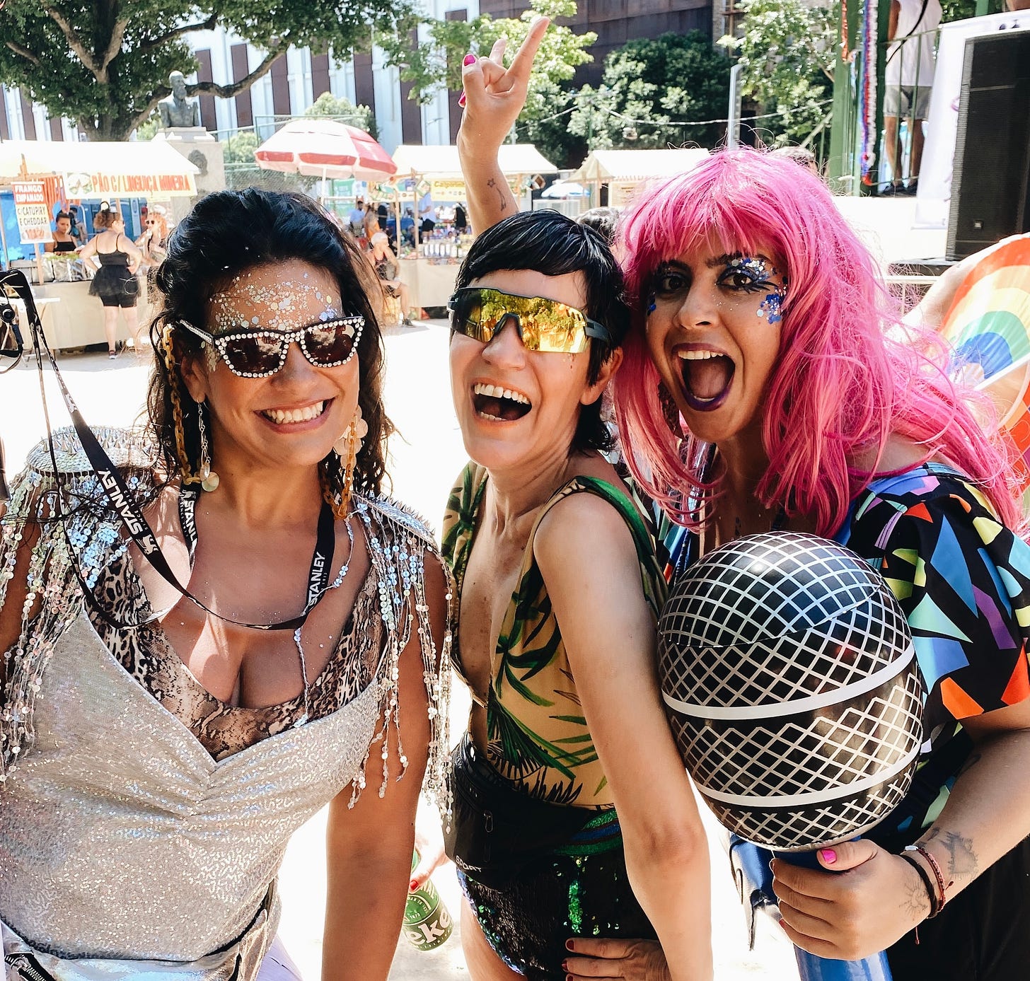 Tres mulheres morenas, uma delas com cabelos longos cor-de-rosa segura um microfone gigante de plástico. Estão maquiadas com glitter e duas estão de óculos escuros. 
