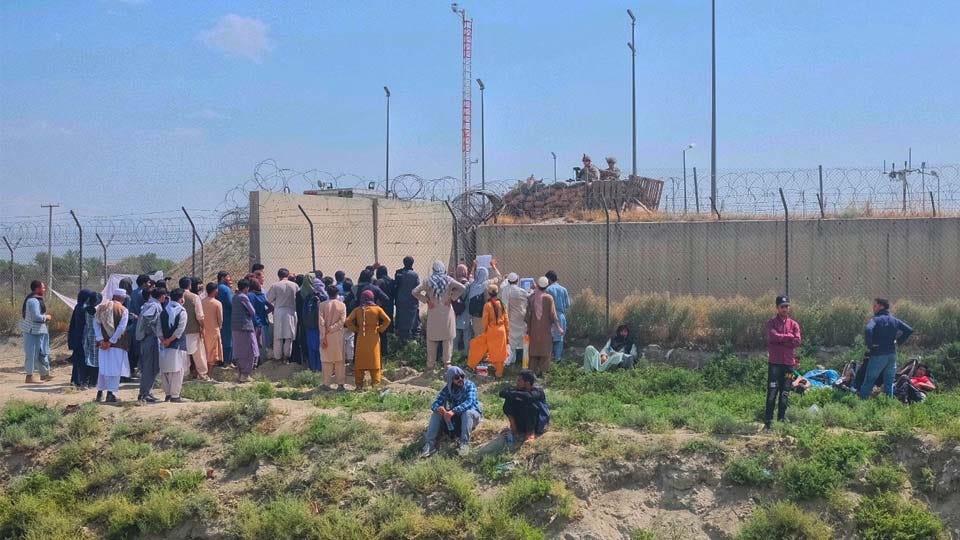 This is a picture showing a group of people wait outside a fence and wall. On the other side are US soldiers. These people await as many evacuate Afghanistan after the Taliban takeover in 2021. 