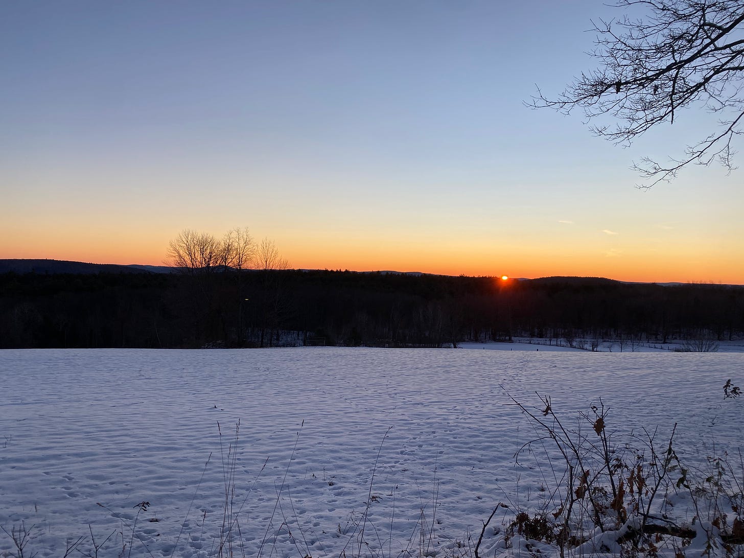 The small golden sun just peeking out from behind a line of dark hills in front of a snowy field. The sky is a clear, pale blue.