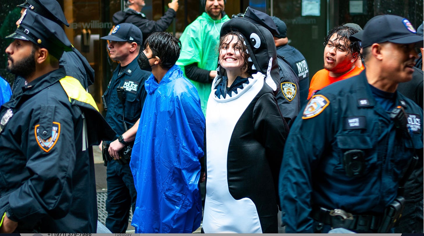 a young white person with hair in their face is in a black and white orca cosute. They are smiling with their flippers behind their back as they are surrounded by NYC police officers