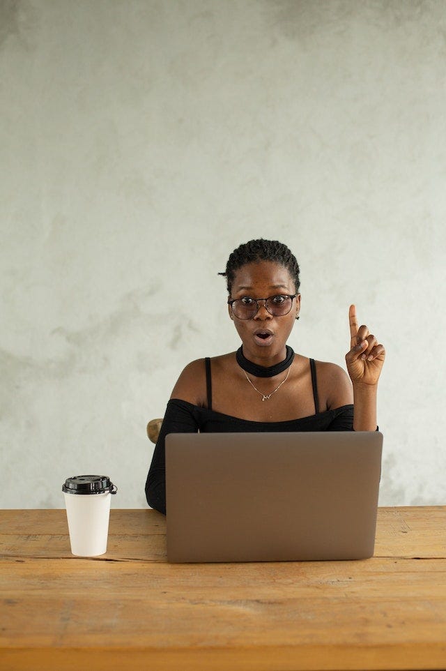 A woman looking surprised at a laptop, with an open mouth and her finger pointing up like she’s just realized something