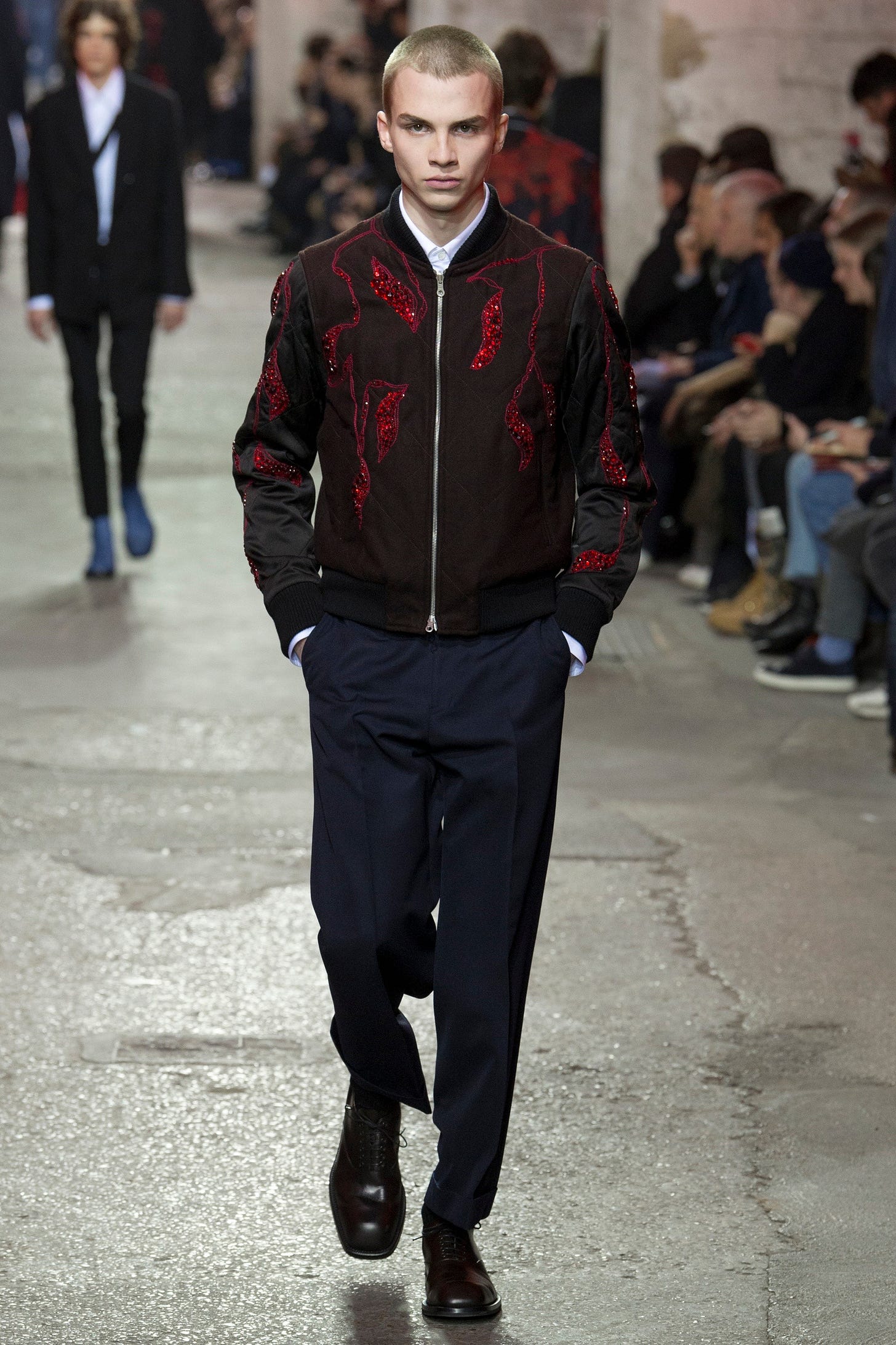 A model on a fashion catwalk, who is a white man with cropped hair, wearing a black bomber jacket with elaborate red embroidery and sequins, navy trousers and formal black shoes. A model on a fashion catwalk, who is a white man with cropped hair, wearing a black bomber jacket with elaborate red embroidery and sequins, navy trousers and formal black shoes.
