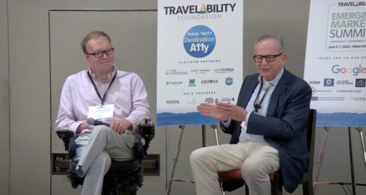 John seated in his wheelchair onstage next to Jake Steinman at the TravelAbility Summit.
