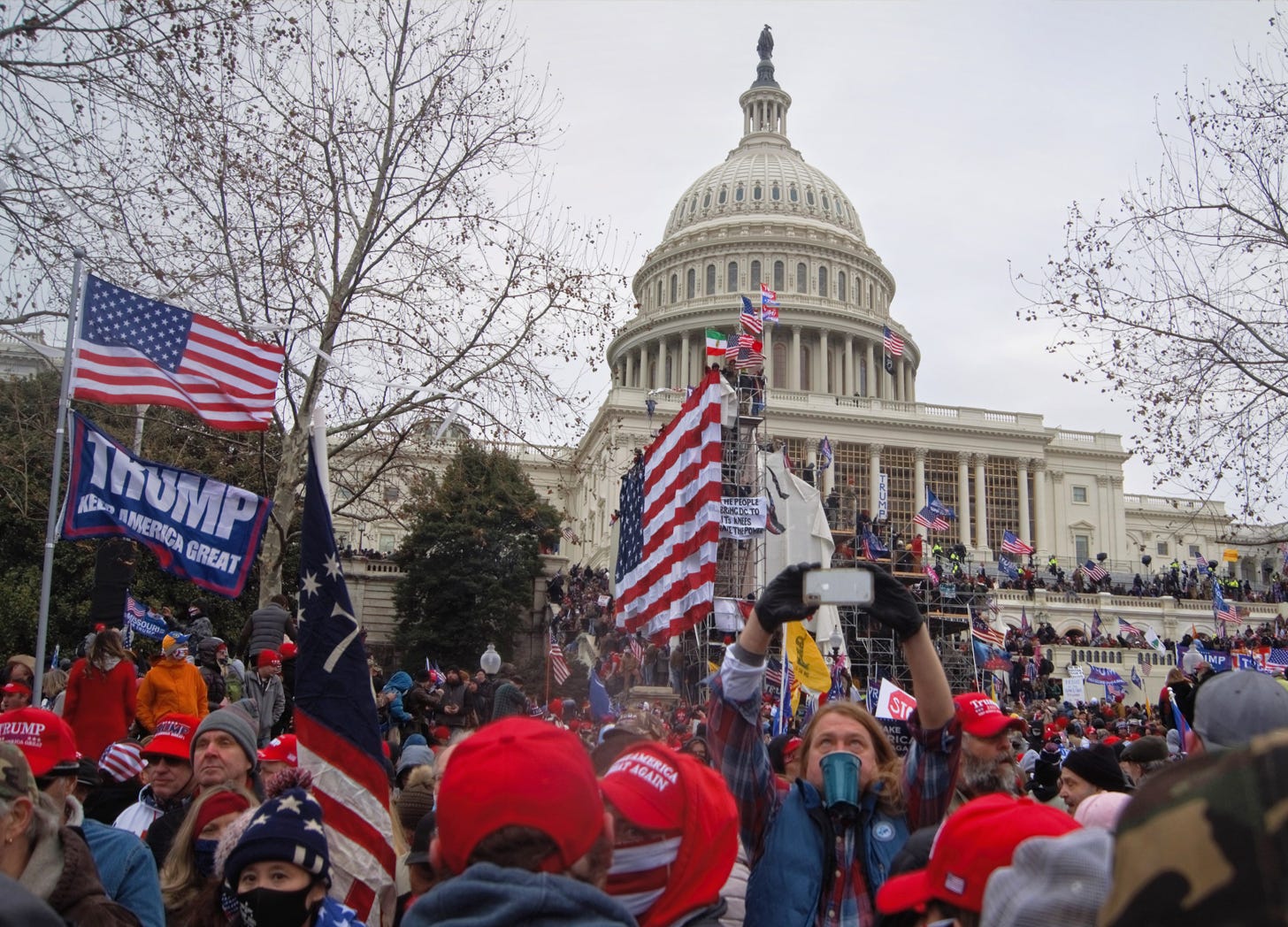 A photo of the January 6, 2021, insurrection at the U.S. Capitol features the Capitol building in the background and a crowd of protesters with American flags and Trump flags in the foreground.