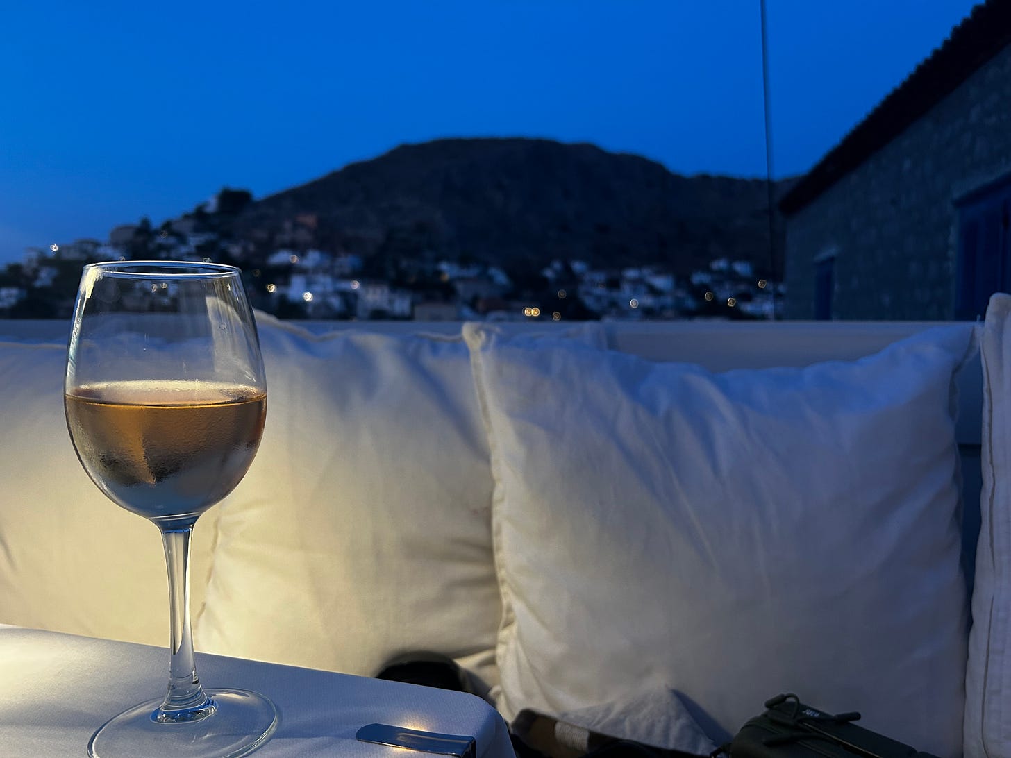 A glass of rosé sits on a table at a restaurant on Hydra, Greece. You can see the town in the background.