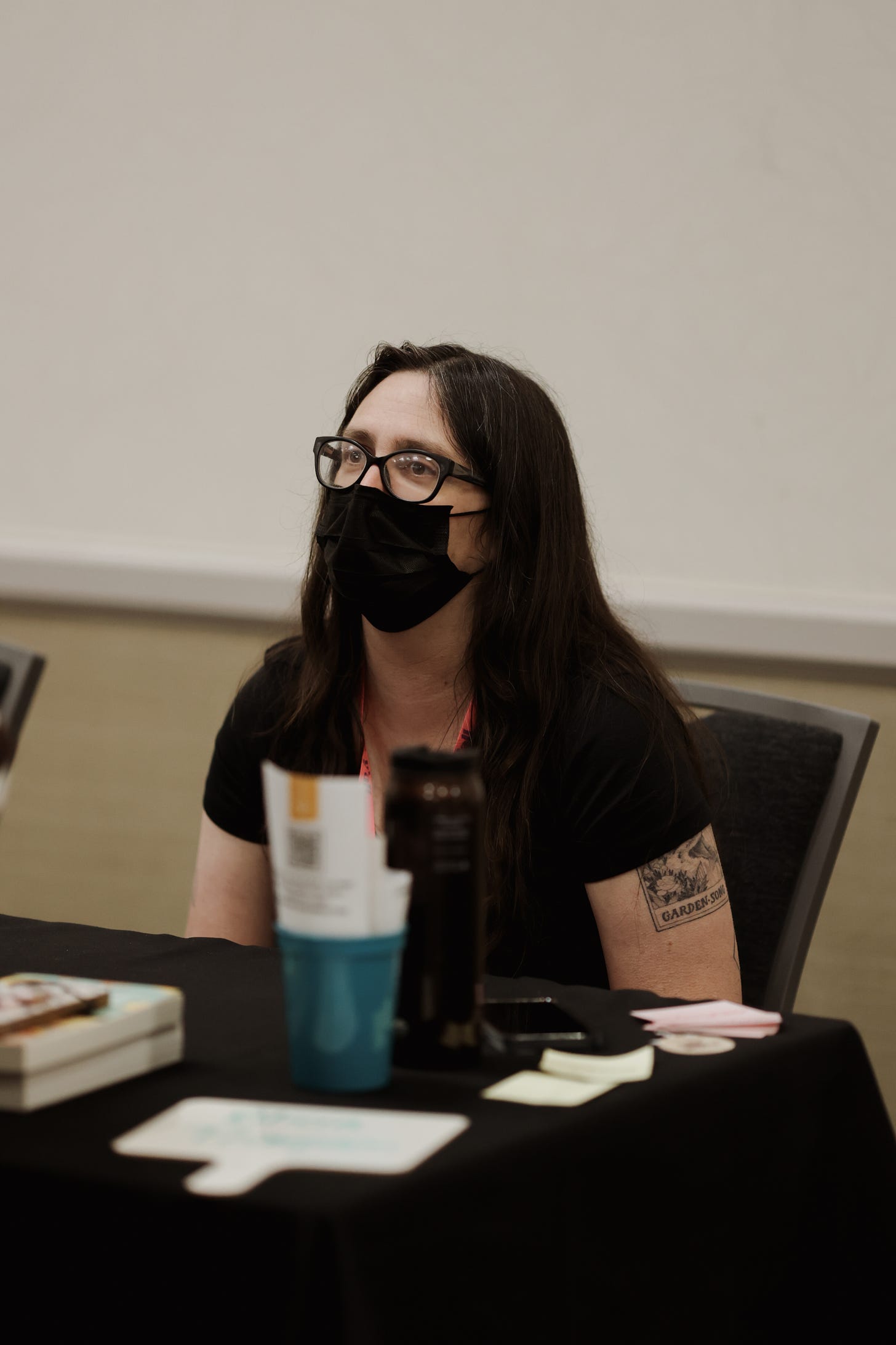 A photo taken by Lauren Wright Payne of me, a white woman wearing a black mask and black glasses, behind a table listening to what someone is saying.