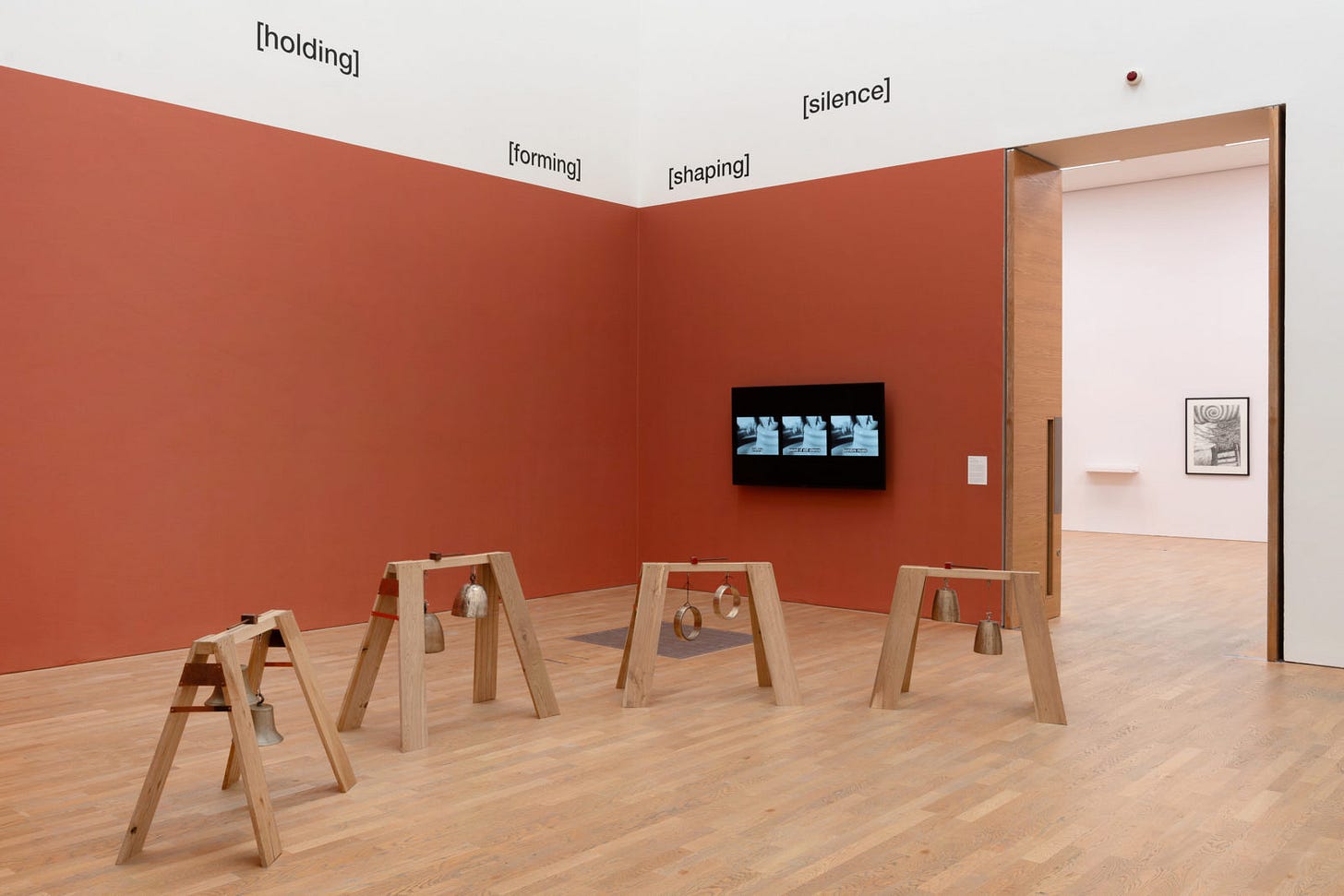 Gallery view of teracotta walls with video installation and four wooden structures holding several metal bells.