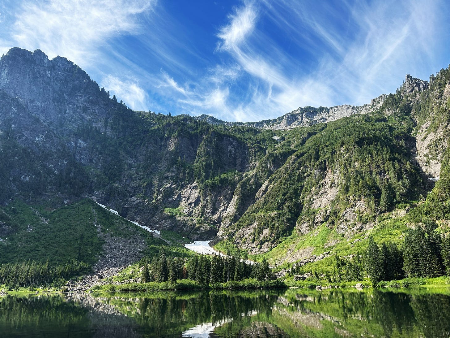 A view of Heather Lake with mountain hills in the background, bright sunshine and wispy white clouds.