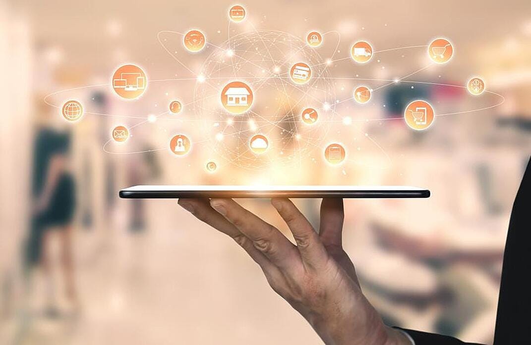 A photograph of an arm carrying a tablet as if it was a drinks tray and they are a waiter. On top of the tablet are icons and graphics connected by several elliptical lines, implying an Internet of things Connectivity being served up.