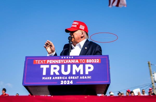 A person in a red hat and a blue suit speaks at a lectern. A red oval is drawn around what appears to be a bullet’s path.