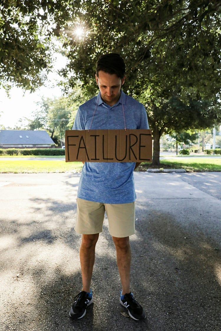 A man in a blue shirt and short trousers holding a brown cardboard placard with the word “FAILURE” in capital letters.