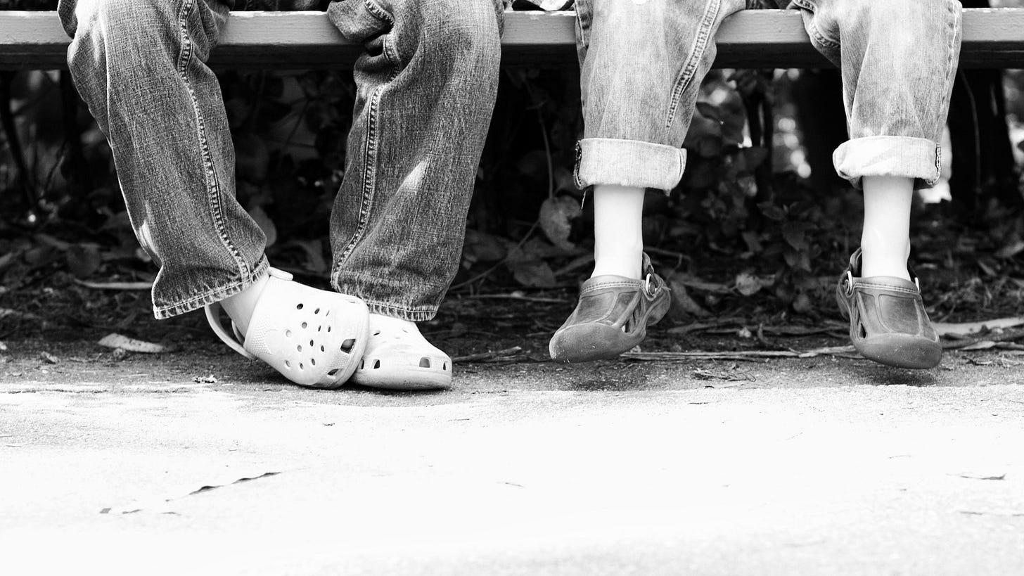 Two pair of feet in crocs rubber shoes of boys sitting on a bench 