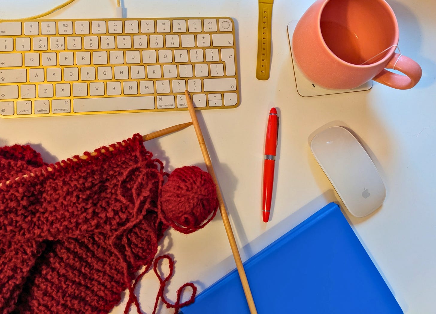An image that looks down on a desk with a keyboard, mouse, pen, notebook and knitting needles and a red ball of wool. There is a pink mug too.