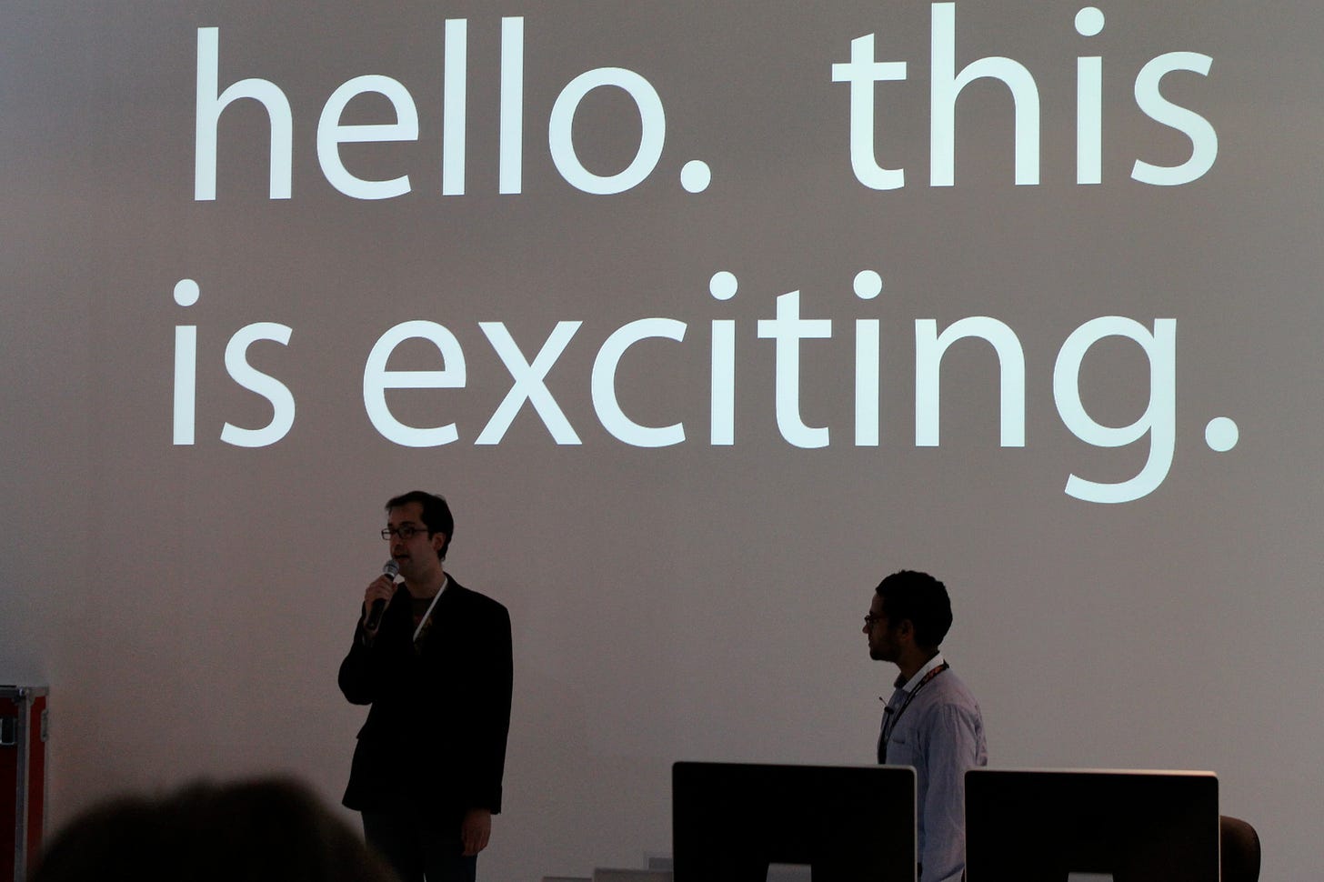 A photo of two people standing in front of a large screen projecting the words: "hello. this is exciting."