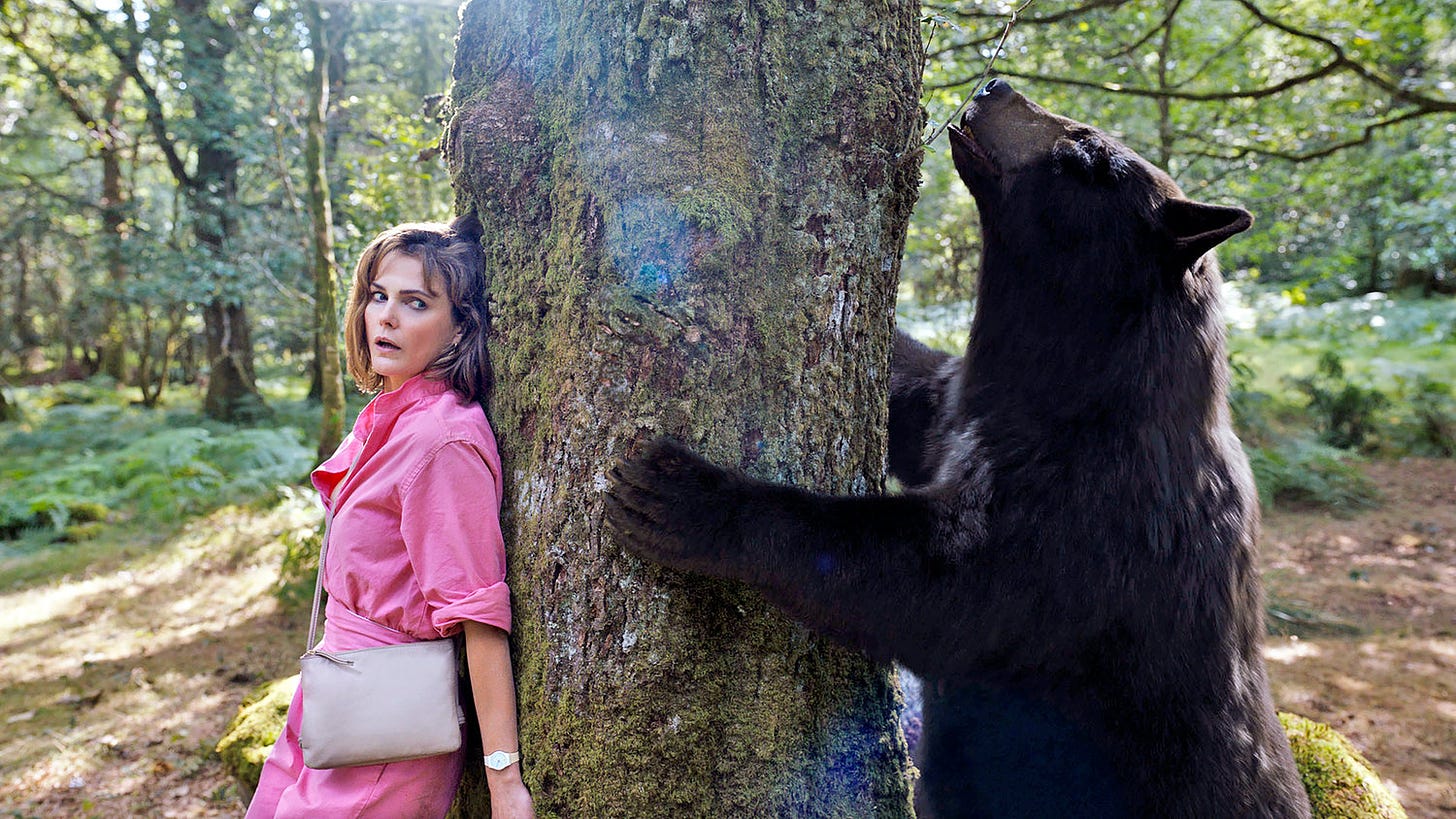 Keri Russel hides on the opposite side of a tree from the Cocaine Bear searching for her in the forest.