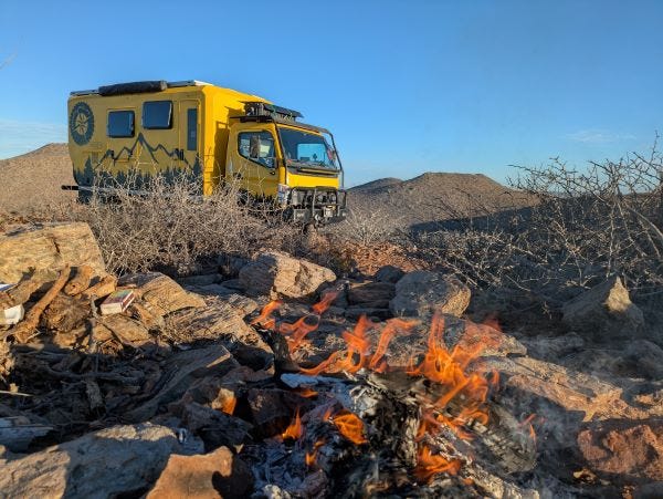 our big yellow truck, Walter, amidst desert plants near a campfire, with the long rays of warm sunrise bathing everything in beautiful light