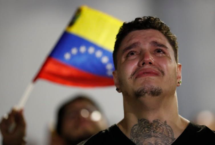 A tattooed Venezuelan man crying with someone waving a Venezuela flag behind him.