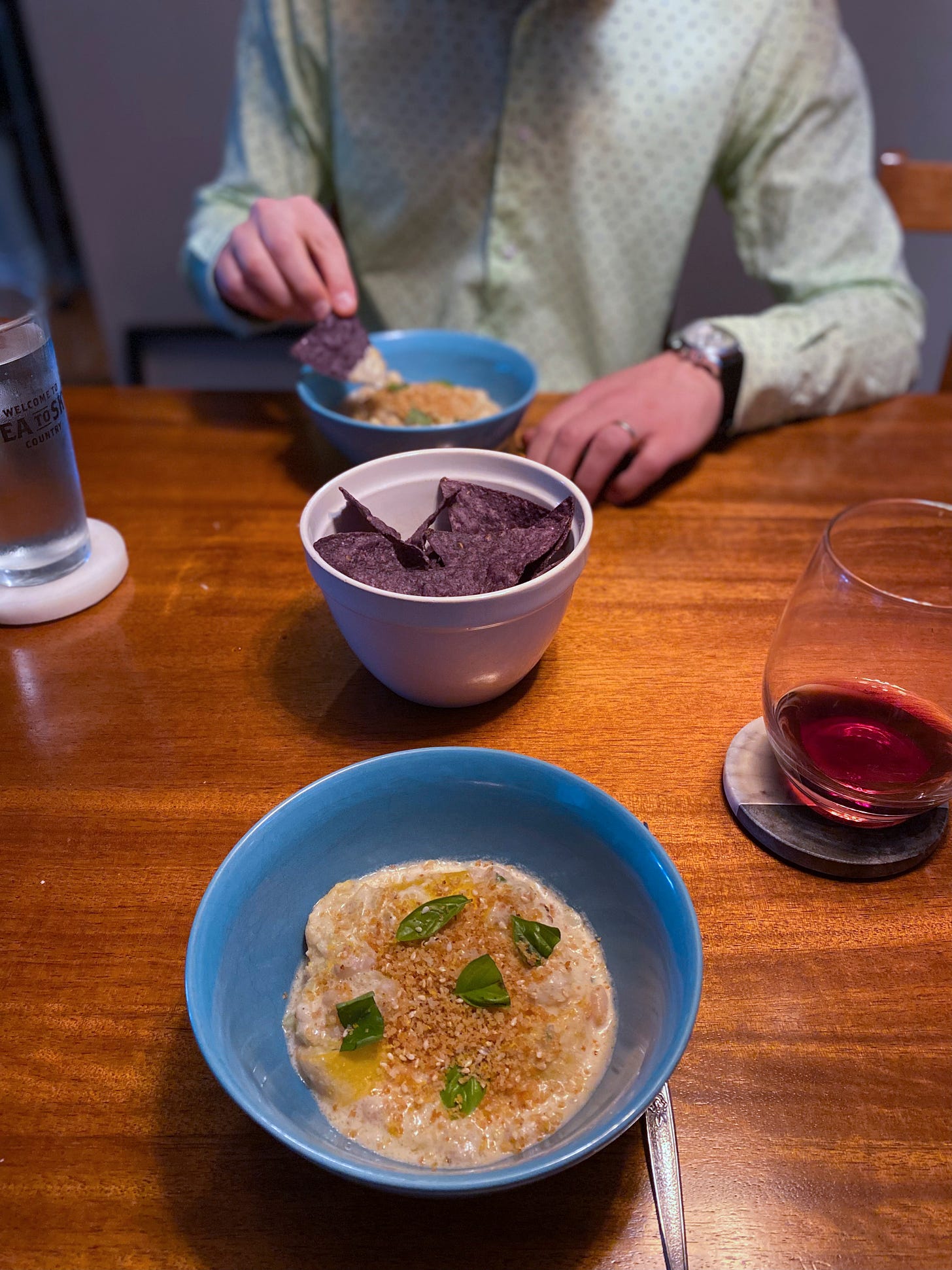 Two blue bowls of the dish described above, topped with panko crumbs and torn basil. In between them is a white bowl of blue corn tortilla chips. Jeff is dipping a chip into his bowl in the background.