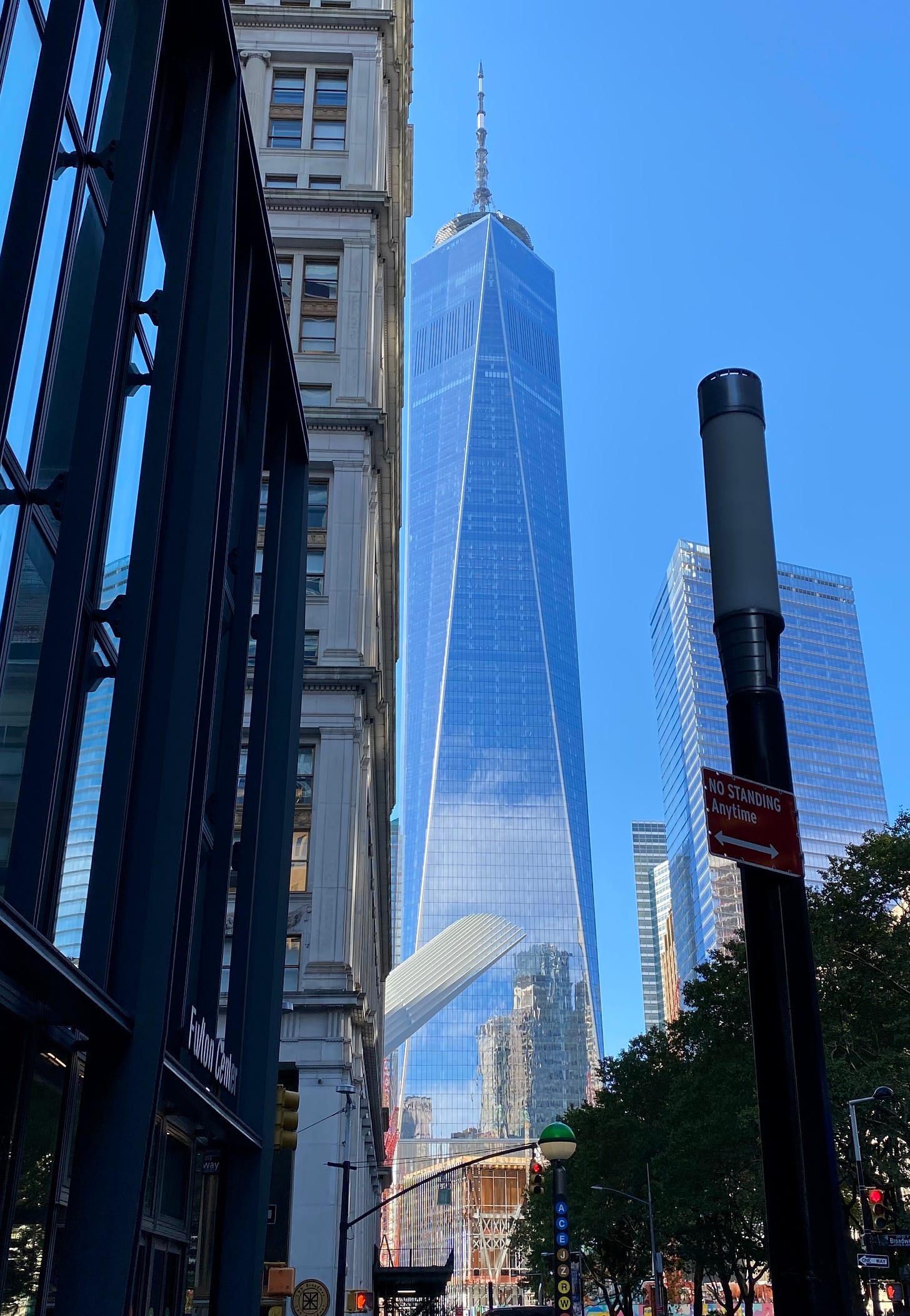 One World Trade Center against a clear blue sky. The light is so bright, you can see the buildings across the street reflected in the its glass facade.