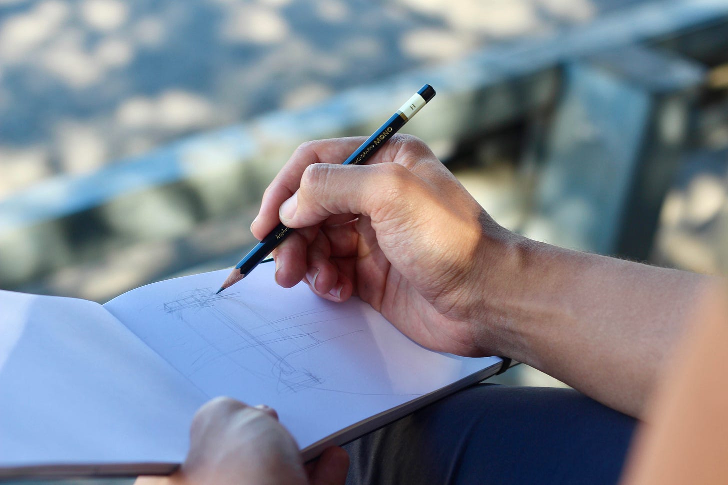 foto no close em um caderno com a mao de um homem branco com um lapis fazendo um esboço em seu caderno, sentado ao ar livre