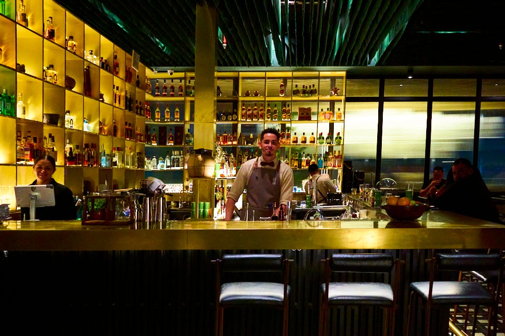 Bartender at Maito in front of a well-lit bar with shelves of Japanese liquor and Pisco greeting patrons. 