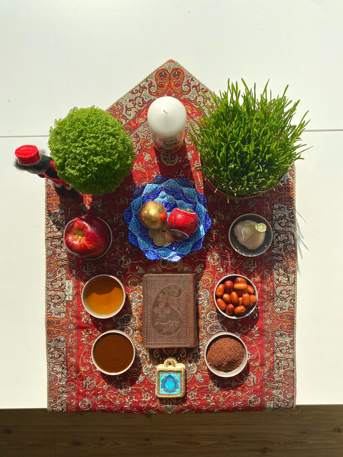 Items for the 7 s table on a beautiful Iranian red patterned cloth, there are herbs, growing wheat, garlic, apple, gold coins, decorated eggs, sumac , candles in decorated ceramic bowls