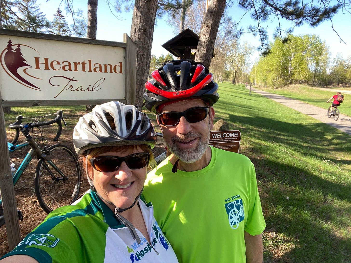 My husband and me, wearing bike helmets and clothing, with bikes in background by sign for the Heartland Trail.