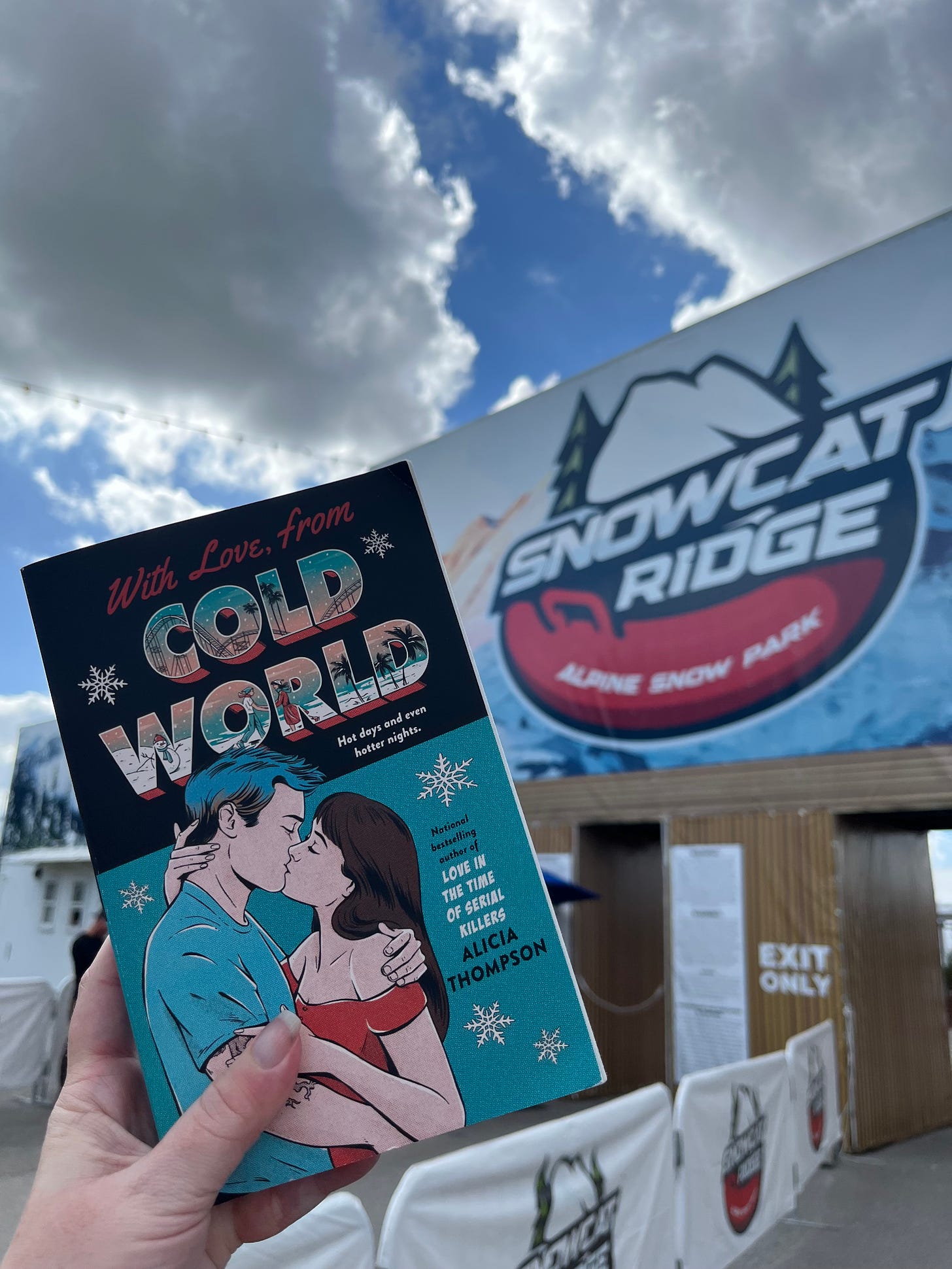 Picture of my hand holding up the teal cover copy of WITH LOVE, FROM COLD WORLD in front of the entrance to Snowcat Ridge Alpine Snow Park