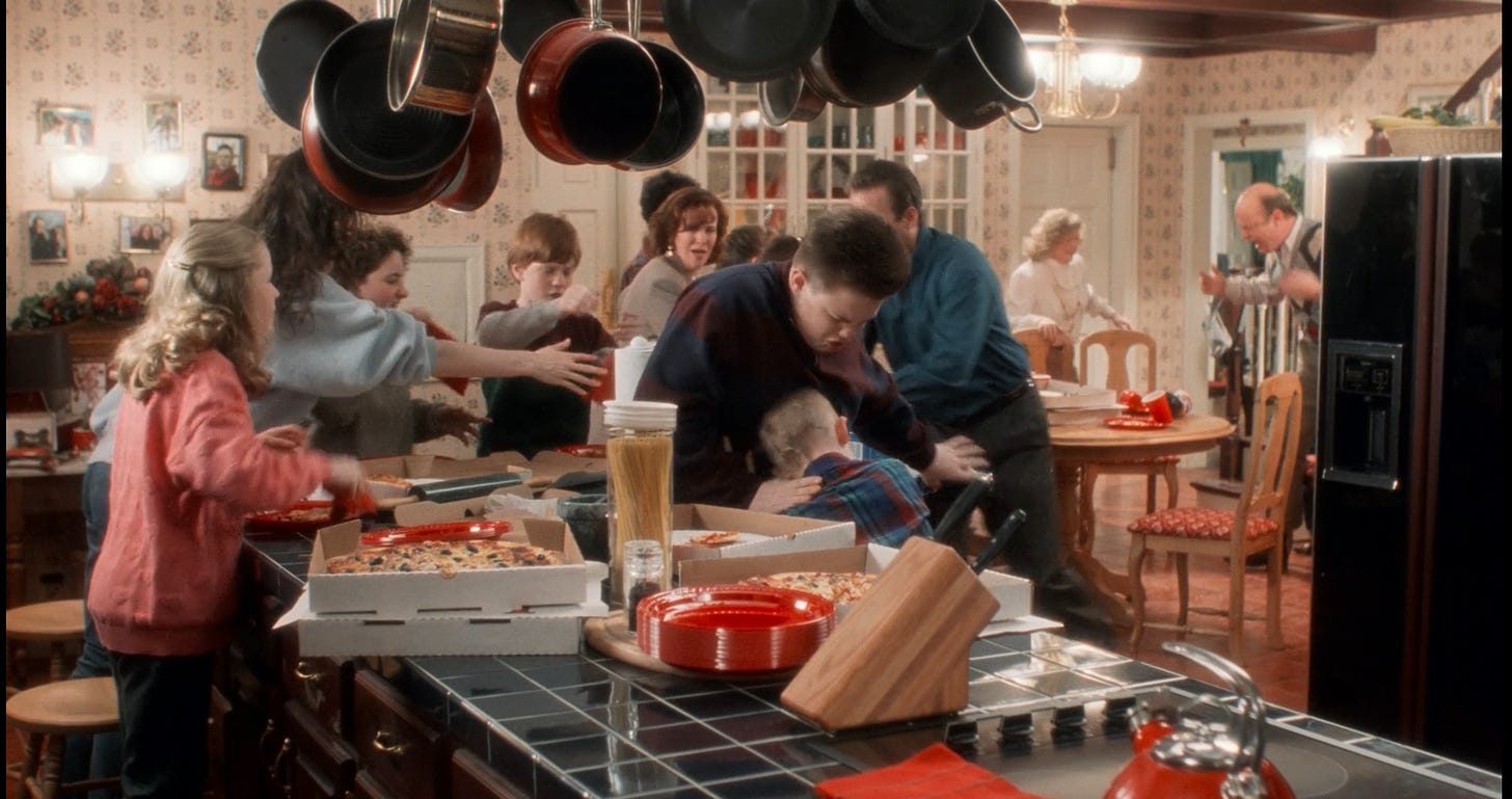 Screenshot of the scene where Kevin is punching Buzz after Buzz ate his pizza. The kitchen has wallpaper, photographs on the wall, hanging pots, and a deep blue tile countertop. There's wood furniture and a very 80s looking black fridge.