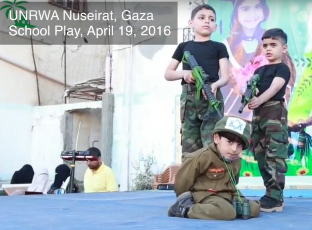 Kids at UNRWA summer camps pointing fake guns at child with Israeli flag on helmet