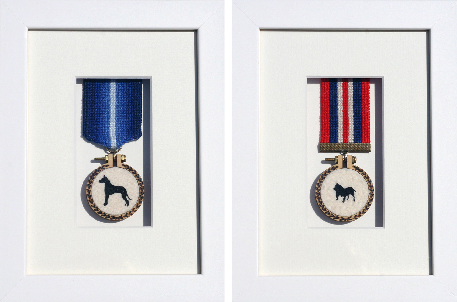 image is of an two framed embroideries side by side in white box frame. Both pieces are embroidered medals, the one on the left has a cross stitched ribbon which has a blue ombré effect going from dark blue either side to light in the middle. Hanging from this is a wooden mini hoop with a black silhouette of a Great Dane stitched on cream cotton. The wooden hoop frame has pyrography detail showing a laurel reef pattern. The medal on the right is similar but has a red white and blue striped cross stitch ribbon and the silhouette of the dog is that of a British bulldog.