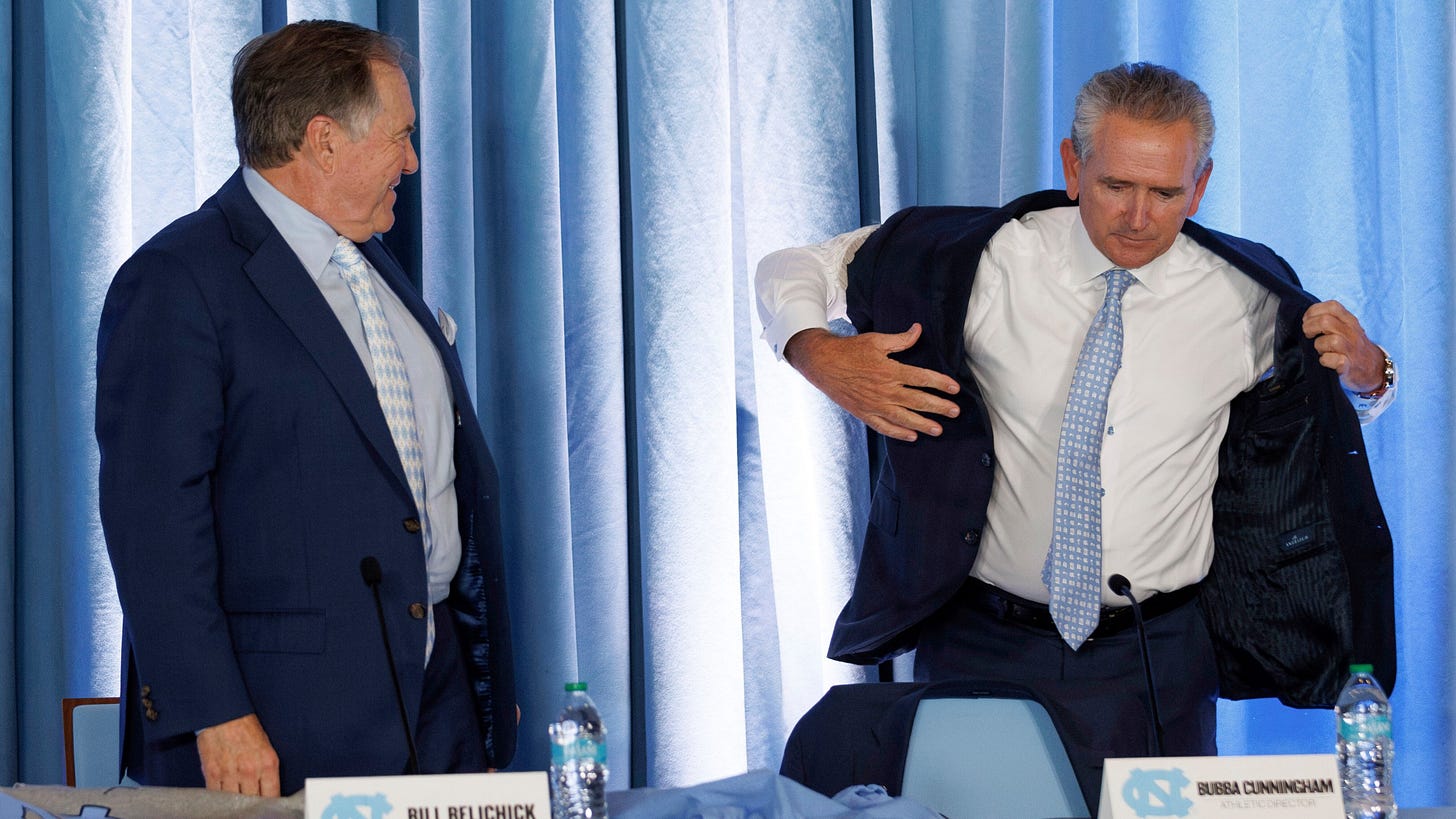 New North Carolina football coach Bill Belichick, left, looks on as UNC athletic director Bubba Cunningham, right, puts on a sport coat with cut-off sleeves during an NCAA college football press conference, Thursday, Dec. 12, 2024, in Chapel Hill, N.C. (AP Photo/Ben McKeown)