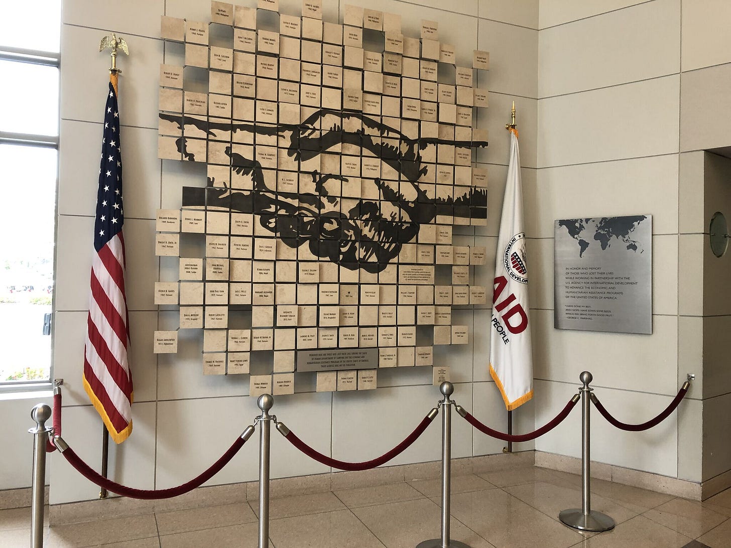 Small plaques with names align with the image of USAID's logo featuring two hands shaking. The American and USAID flags border the memorial