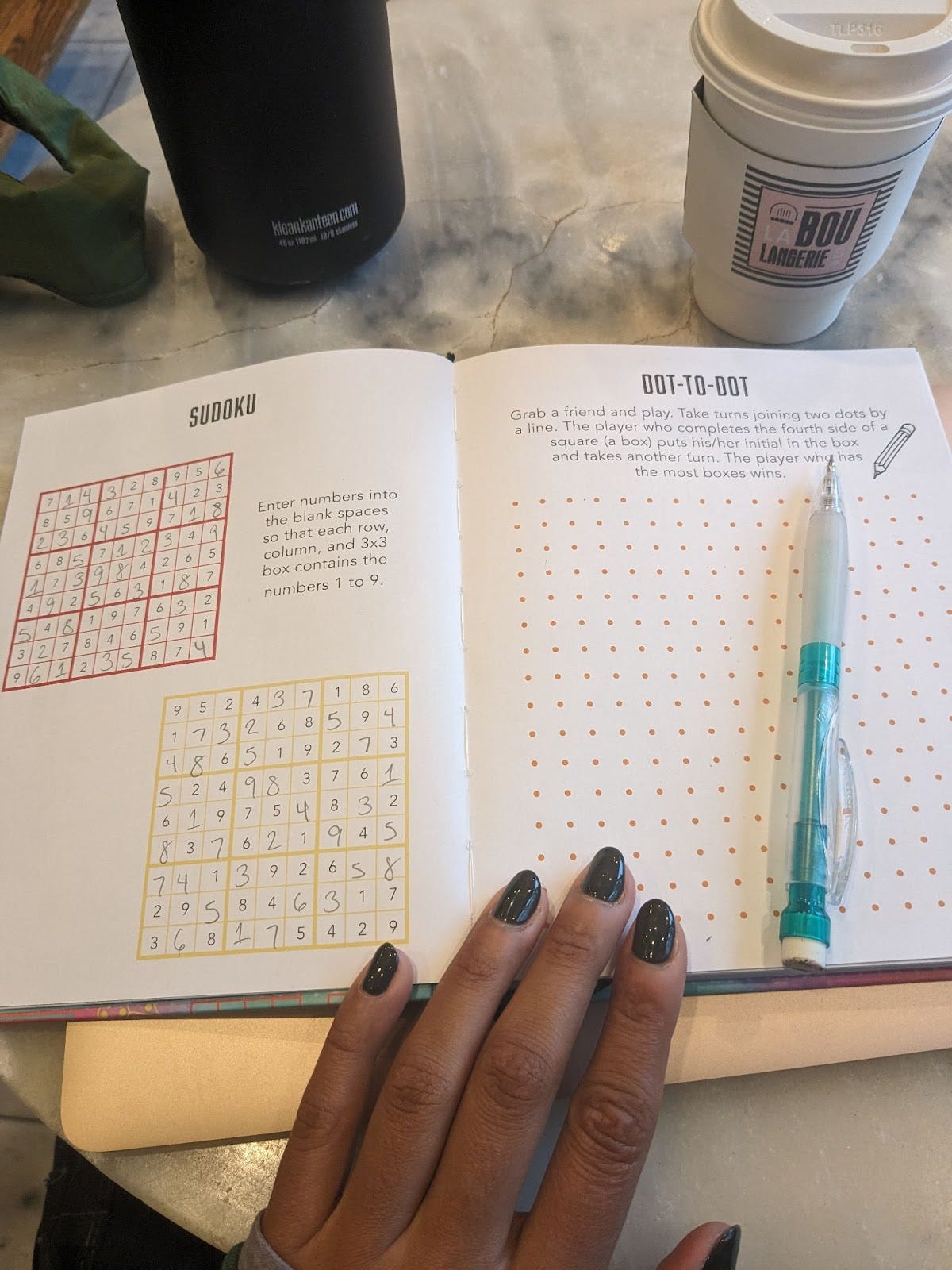 A book with the page turned to 2 sudoku puzzles. A coffee cup that says "boulangerie" on it is by the book.
