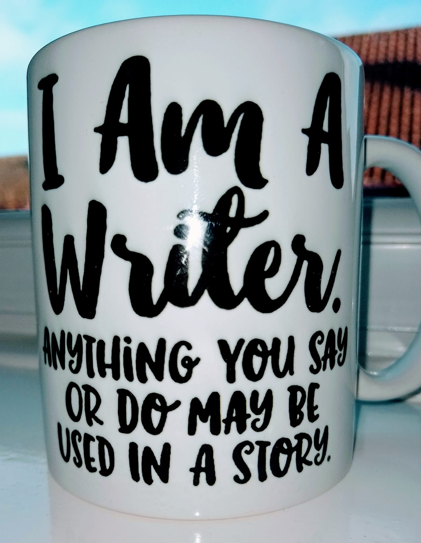 A photograph of a white tea mug sat on a window shelf, decorated wth black text stating: "I am a writer. Anything you say or do may be used in a story."