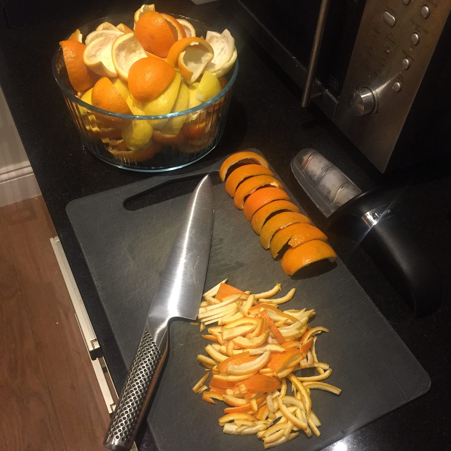 A glass bowl of quartered orange and lemon peel sits on a black workshop next to a chopping board, global knife, knife sharpener and a pile of finely cut peel