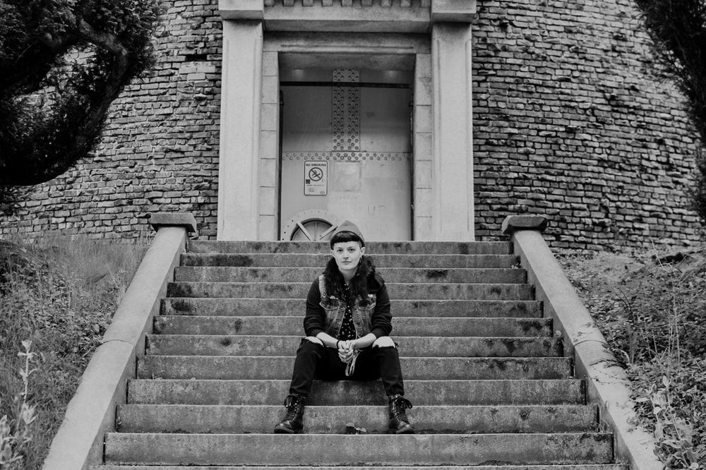 B+W photo of Dana Winter (they/them) sitting on concrete stairs in front of a brick water tower
