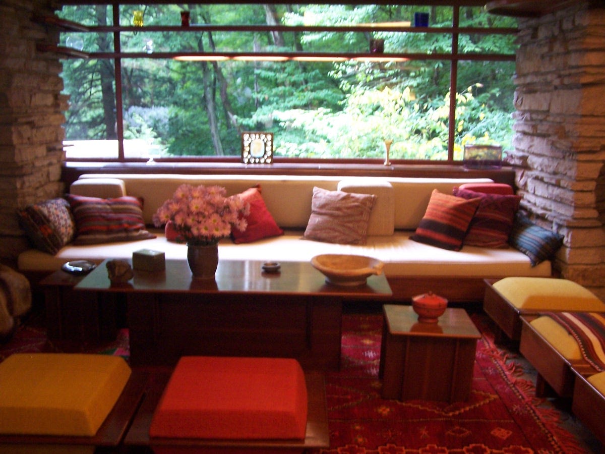 A long, low couch against a wall of windows framed with stone walls, looking out into a forest. Cushioned stools and coffee tables with decorative bowls sit in front of the couch.
