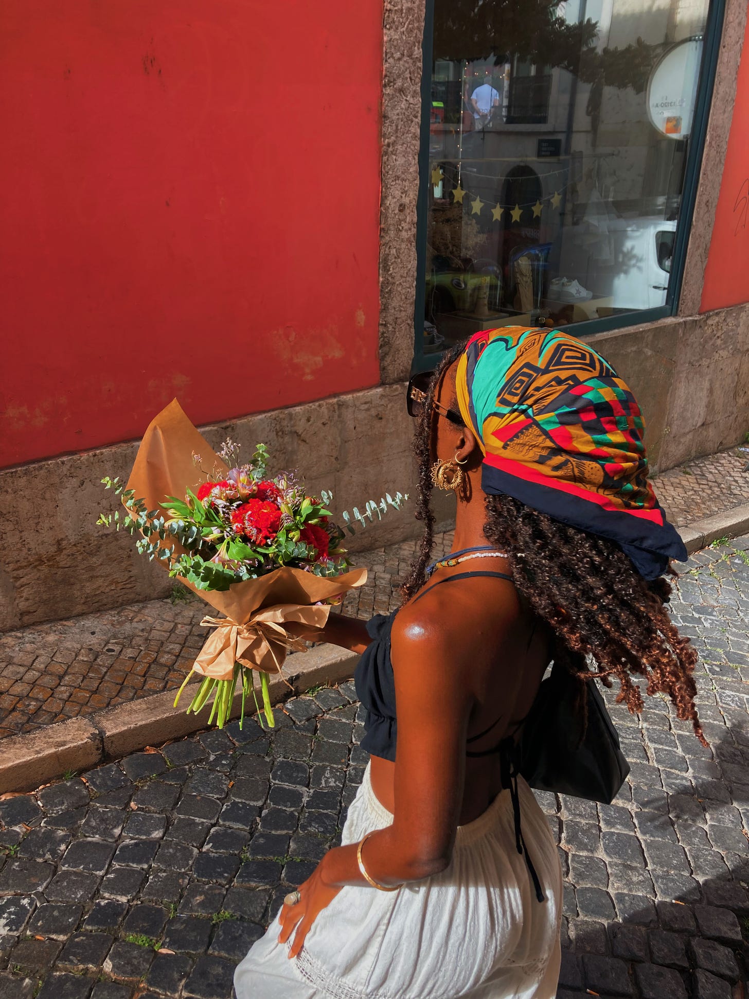 Amara wearing a colourful headscarf caught mid-spin in front of a red wall in Lisbon. She is wearing a white maxi skirt and a black top and holding a red and purple bouquet with eucalyptus stems too. She is wearing all of her favourite gold earrings and crystal necklaces.